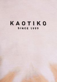 Světle růžové tričko s tie-dye vzorem a černým logem "KAOTIKO" a textem "SINCE 1999", má měkkou bavlněnou strukturu a volný střih.