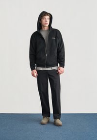 Sweat à capuche zippé en polaire noire avec capuche, t-shirt gris en dessous, pantalon noir et chaussures beige clair, porté sur un fond clair.