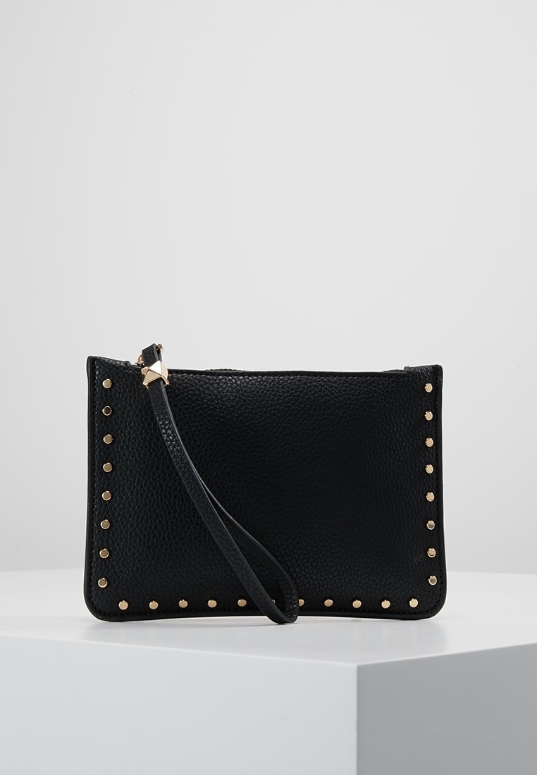 Pochette bracelet texturée noire avec des clous dorés le long des bords et une fine lanière attachée près de la fermeture éclair, présentée sur un piédestal blanc.