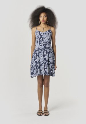 Marineblauwe bloemenjurk met blauw-witte patronen; nauwsluitend lijfje, uitlopende rok, spaghetti bandjes. Gecombineerd met zwarte sandalen, staat model rechtop.