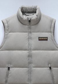 Ljusgrå ärmlös puffer-väst med hög krage, vertikal quiltning och en zippered ficka, med en märkespatch på vänster sida.