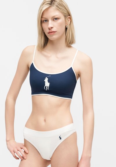 Νεαρή γυναίκα που φοράει μπλε σκούρο crop top και λευκά μαγιό μπικίνι κάτω μέρος με λογότυπο παίκτη πόλο αλόγου σε όλα τα ρούχα.