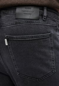 Κοντινό πλάνο πίσω τσέπης μαύρου τζιν με δερμάτινη ετικέτα Mads Nørgaard Copenhagen και λευκή υφασμάτινη ετικέτα με το λογότυπο.