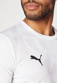 Hvid atletisk trøje med et subtilt mønster og et sort Puma-logo på brystet. Lavet af glat, let materiale.