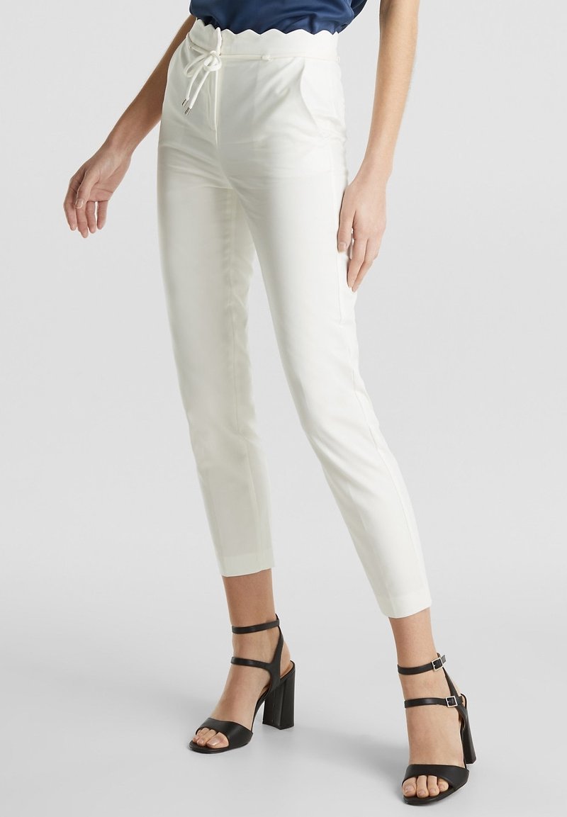 Femme portant un pantalon blanc taille haute et fuselé avec une ceinture festonnée et des sandales noires à talons larges et à brides, debout sur un fond gris.