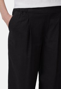Zwarte geplooide broeken met een elastische tailleband. Bevat twee zijzakken en een textuurstof. Close-up van de tailleband en het zakgebied.