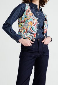 En denimskjorta under en färgglad mönstrad väst. Västet har svallande mönster i blått, orange och grönt. Mörka jeans med lapplommer.