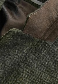 Wollstoff hat eine strukturierte grüne Außenfläche und ein glattes braunes Innenfutter. Das Detail umfasst sichtbare Nähte entlang der Kanten.