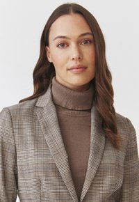 Hnědý károvaný blazer se strukturovanými klopami, nošený přes přiléhavý hnědý rolák. Hladký textil s jemnými vzorovými detaily.
