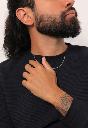 Silberne gedrehte Kettenhalskette, die über einem strukturierten dunklen Hemd getragen wird. Nahaufnahme einer Hand mit sichtbarem Tattoo, die leicht auf der Brust ruht.