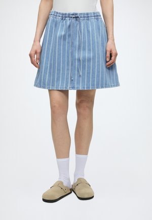 Femme portant une jupe en denim à rayures verticales bleues et blanches avec une taille élastique et un cordon, des chaussettes blanches et des chaussures beige à enfiler.