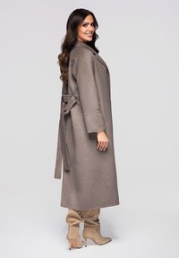 Lange, lichtbruine jas gemaakt van zacht materiaal met een ceintuurstrik in de taille, brede mouwen en een knielang ontwerp, gecombineerd met beige puntige laarzen.