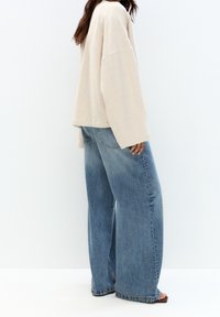 Beige oversized longslavenshirt, gecombineerd met losse lichtblauwe denim jeans. Getextureerde stof en een relaxed fit met een wijduitlopende snit.