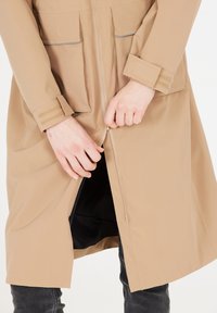 Beige zip-up frakke med to frontlommer, lange ærmer med velcrobånd og todelt lynlåsdetalje, vist delvist lynlåst.