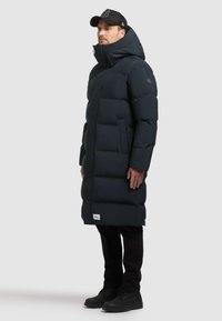 Tumesinine tikk-jahtud jääkapp kõrge kaelusega, lukuga ja isoleeritud disainiga. Omab küljetaskuid ja logopatchi allääres. Mustad püksid ja saapad.