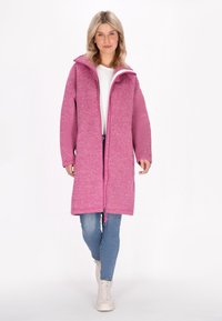 Lungo cappotto rosa con finitura testurizzata, zip frontale e cordino. Indossato sopra una camicia bianca e jeans blu, abbinato a sneaker di colore chiaro.