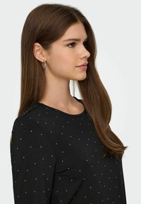 Maglietta nera a manica lunga con scollatura rotonda, caratterizzata da borchie argentate sparse. Tessuto liscio e vestibilità aderente. Volto mostrato di profilo.