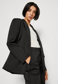 Black tailored blazer with a satin lapel, structured shoulders, and a slim fit. Worn over a white textured top, featuring a pocket.