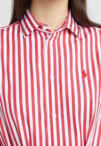 Czerwono-biała koszula z guzikiem w paski, z wyrazistym kołnierzykiem i haftowanym logo na piersi. Materiał wydaje się gładki i lekki.
