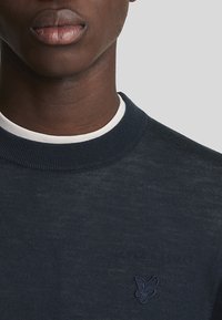 Pullover in maglia blu navy con colluccio a coste. Presenta un ricamo logo discreto sul petto. Tessuto leggero e design aderente.