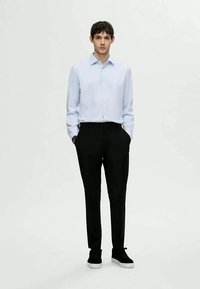 Camisa de manga longa azul clara com colarinho pontudo, combinada com calças pretas e ténis de sola branca, de pé diante de um fundo neutro.