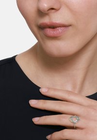 Anello d'argento con un design a raggi di sole, caratterizzato da una pietra centrale, appoggiato su una camicia nera. Il tono della pelle è visibile sulle dita e sul collo.