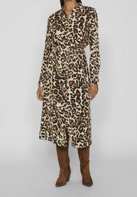 Vestido de manga larga con estampado de leopardo en beige, negro y marrón, con cuello y un cinturón de lazada en la cintura. Combinado con botas marrones hasta la rodilla.