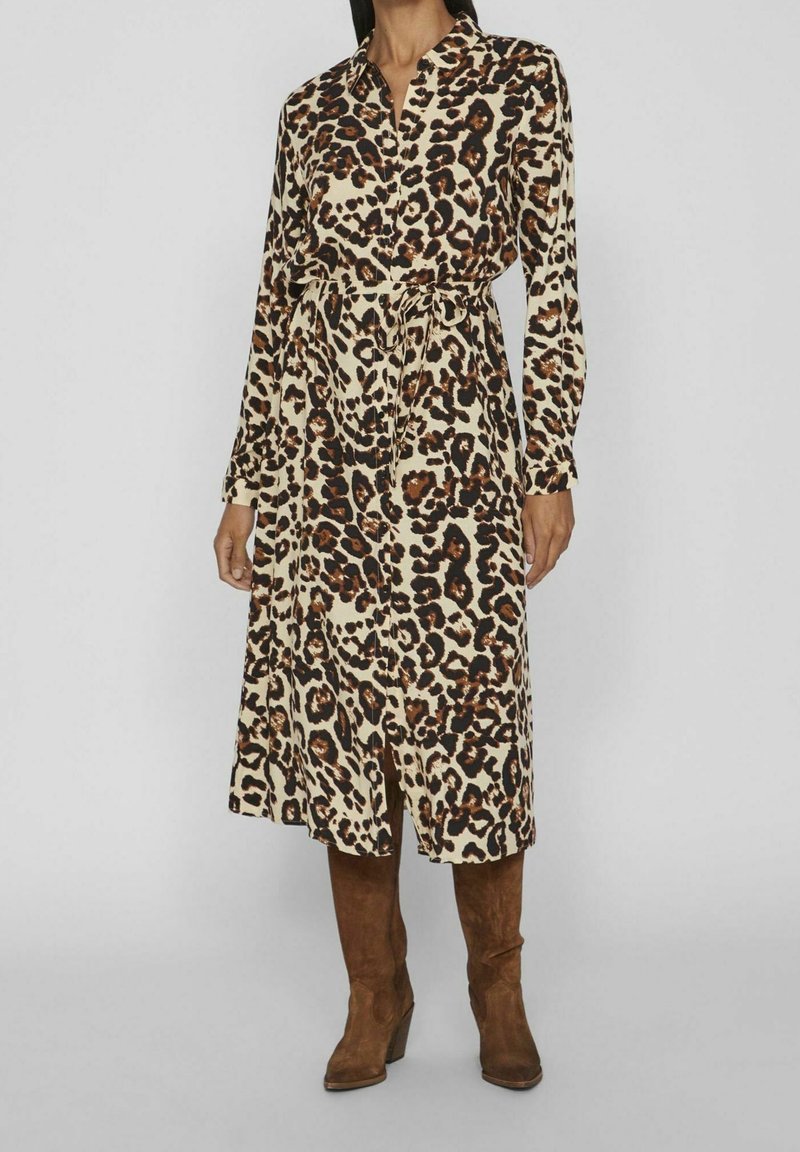 Vestido de manga larga con estampado de leopardo en beige, negro y marrón, con cuello y un cinturón de lazada en la cintura. Combinado con botas marrones hasta la rodilla.