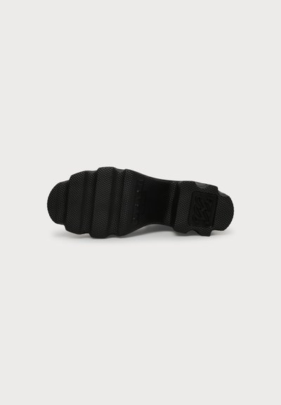 Semelle de chaussure texturée noire avec motif de grip côtelé et un petit logo embossé près du talon sur fond blanc.