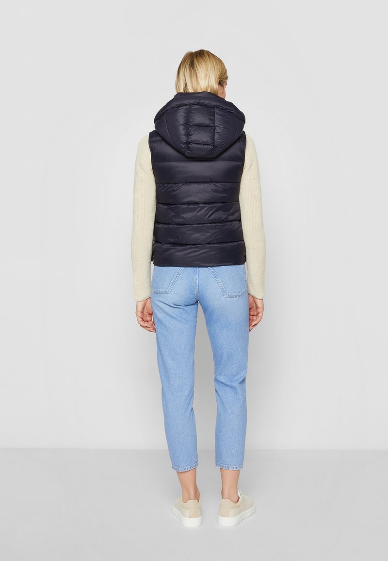 Eine Frau von hinten, die eine marineblaue Steppweste mit Kapuze, einen beigen Pullover, hellblaue, verkürzte Jeans und weiße Sneaker auf einem schlichten Hintergrund trägt.