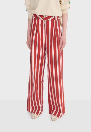 Røde og hvide bukser med lodrette striber og brede ben, båret med beige top og cremefarvede spidse sko mod en lys baggrund.