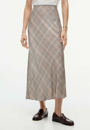 Donna che indossa una gonna midi a quadri beige e neri, top bianco, orologio da polso dorato e mocassini in pelle marrone scuro, in piedi su un pavimento bianco.