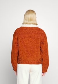 Hnědo-oranžová kordová bunda s huňatým bílým kožešinovým límcem, s texturovaným vzorem, oversized střihem a uzávěry na patentky.