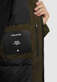 Ruka drži otvorenú olivovo zelenú bundu, na ktorej je viditeľná vnútorná nášivka s nápisom "KhujoTM Genuine Brand Established 2001" a zipsová vnútorná vrecko.