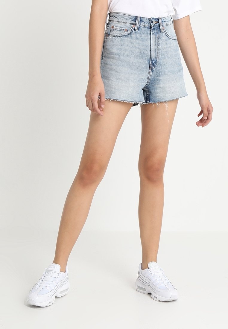 Personne portant un short en jean bleu clair effiloché, des baskets blanches et un haut blanc, debout devant un fond blanc uni.