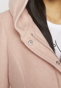 Giacca cappuccio rosa in tessuto texturizzato, con chiusura a zip argentata e bottoni a pressione. All'interno si vede una t-shirt grafica bianca.