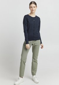 Cardigan en maille bleu marine avec un col rond, fermeture à boutons sur le devant et manches longues, associé à un pantalon longueur cheville vert clair et des baskets blanches.