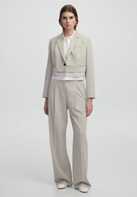 Blazer cropped beige con un singolo bottone, abbinato a pantaloni beige larghi. Camicia bianca sotto e scarpe bianche con texture.