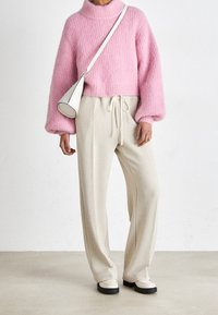 Roze gebreide trui met een hoge hals en ballonmouwen, gecombineerd met beige gestructureerde broeken en een witte handtas met een ronde basis.