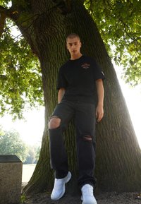 Black cotton t-shirt with logo, distressed black denim jeans with rips, and white athletic shoes, leaning against a large tree trunk.