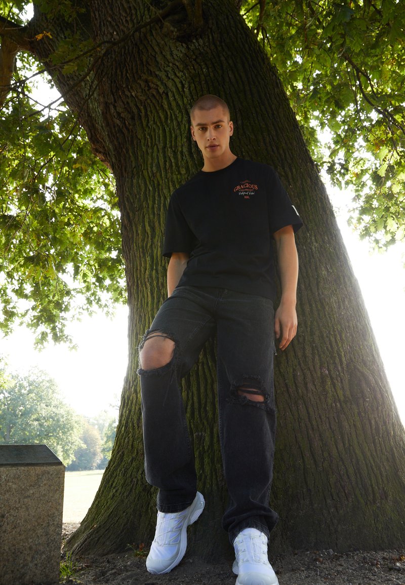 Black cotton t-shirt with logo, distressed black denim jeans with rips, and white athletic shoes, leaning against a large tree trunk.