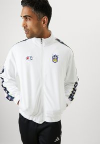 Witte zip-up jas van gladde stof met een logo, contrasterende zwarte en gekleurde afwerking op de mouwen, en elastische manchetten en taille.