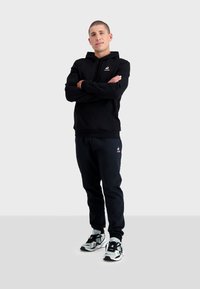 Sudadera negra con capucha y pantalones de chándal a juego en una tela suave, con un pequeño logo en el pecho y en el costado, llevados con zapatillas deportivas negras y blancas.