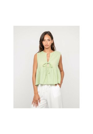 Femme aux longs cheveux bruns portant un haut vert clair sans manches avec un nœud à l'avant et un pantalon blanc ample, debout devant un fond uni.