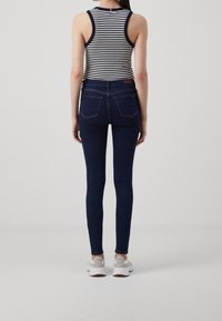 Jeans skinny de mezclilla azul oscuro con cintura alta, bolsillos traseros sutiles, combinados con una blusa sin mangas a rayas blancas y negras y zapatillas blancas.