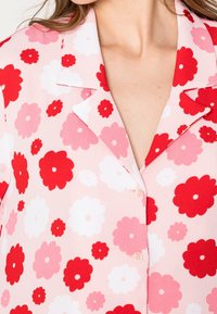 Camicia in tessuto leggero con motivo floreale. Presenta fiori rossi, rosa e bianchi su uno sfondo rosa chiaro. Collo a punte con bottoni.