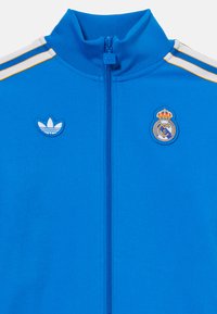 Blå idrettsjakke med høy krage og glidelås, med hvite og gullstriper, adidas- og Real Madrid-logoer, samt en jevn tekstur.