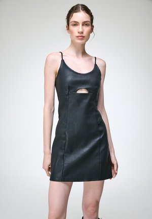 Femme portant une mini robe noire en cuir sans manches avec de fines bretelles et une petite découpe à l'avant, debout devant un fond clair uni.