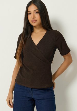 Jeune femme aux longs cheveux raides porte un top cache-cœur à manches courtes marron foncé et un jean bleu foncé, debout devant un fond clair uni.