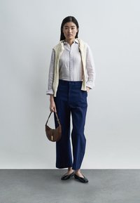 Tânără stând, purtând o cămașă în dungi, un pulover crem așezat peste umeri, pantaloni albaștri, balerini negri, ținând o geantă maro într-o cameră albă.